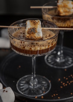 A close-up of a cocktail glass filled with a coffee-based beverage, topped with a roasted marshmallow skewer. The drink appears creamy and is garnished with cocoa or coffee powder, suggesting a dessert-like quality. The setting has a cozy, warm ambiance with a dark background that contrasts with the glass and marshmallow.