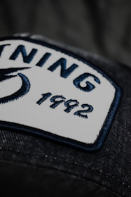 A close-up view of a rectangular white patch with a navy blue border sewn onto a dark fabric. The patch contains two words 'NING' and '1992' embroidered in navy blue thread. Part of a blue design, possibly a logo, is visible on the left side of the patch.