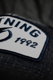 A close-up view of a rectangular white patch with a navy blue border sewn onto a dark fabric. The patch contains two words 'NING' and '1992' embroidered in navy blue thread. Part of a blue design, possibly a logo, is visible on the left side of the patch.
