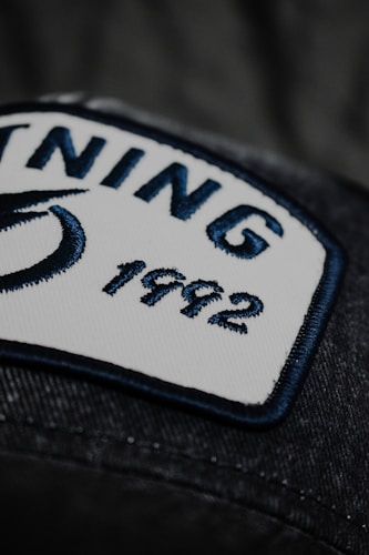 A close-up view of a rectangular white patch with a navy blue border sewn onto a dark fabric. The patch contains two words 'NING' and '1992' embroidered in navy blue thread. Part of a blue design, possibly a logo, is visible on the left side of the patch.