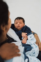 A newborn baby is peacefully asleep, wrapped in a blue and white blanket. The baby is resting in the arms of an adult who is partially visible, suggesting a tender and protective interaction.