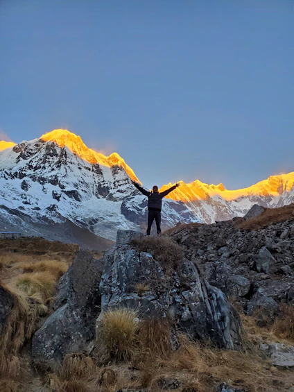 A rugged mountain peak bathed in golden sunrise light, with a lone hiker standing triumphantly.
