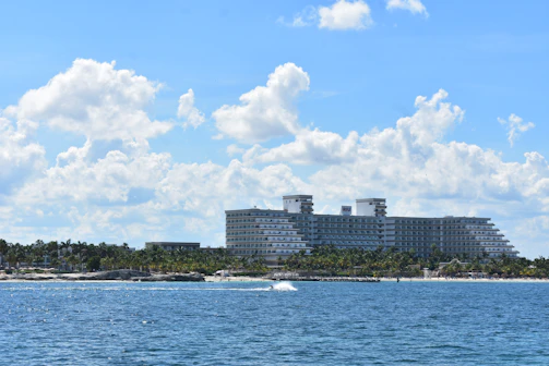 A large, modern hotel building with multiple floors is situated along a shoreline with a partly cloudy sky overhead. The building is surrounded by lush greenery, and the ocean extends into the foreground. A small splash in the water suggests the presence of a boat or jet ski.