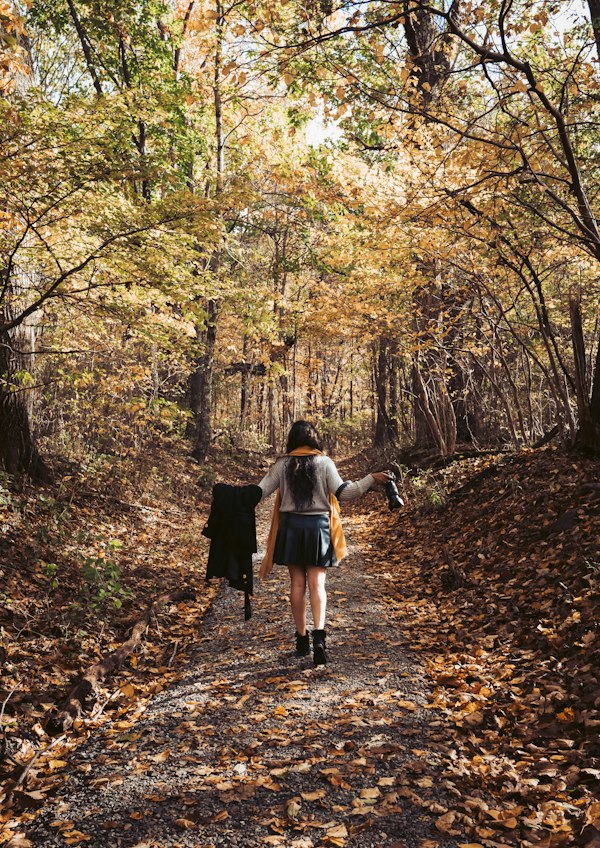 A woman walking alone down a forest path covered in fallen autumn leaves