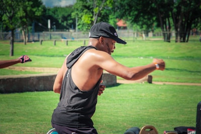 A person wearing a sleeveless black top and cap is engaged in outdoor physical activity, possibly exercising or practicing martial arts, in a grassy park. Trees and park benches are visible in the background, while another person’s arm, wearing a pink glove, can be seen extending into the frame.