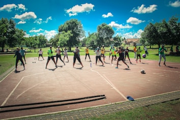 A group of people are exercising outdoors on a concrete court, surrounded by green spaces and trees. They are standing in a formation, appearing to engage in an organized fitness class. Each participant wears athletic clothing, and several carry exercise equipment such as dumbbells and ropes. The sky is bright blue with scattered fluffy clouds, creating a vibrant and energetic atmosphere.