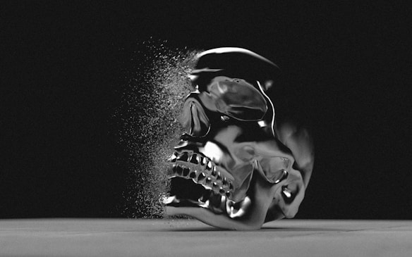 A metallic skull is depicted against a dark background, with part of it breaking apart into small particles, giving the appearance of disintegration. The surface is smooth and reflective, capturing light in a dramatic way.