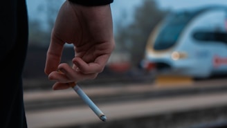 A hand is holding a cigarette between the fingers with a blurred train passing in the background, suggesting a scene at a train station.