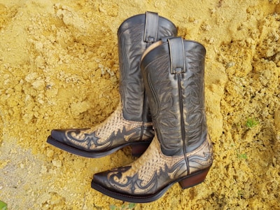 A pair of intricately designed cowboy boots featuring a combination of dark leather and snake skin patterns. They are set against a background of yellowish sand or gravel.