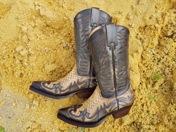 A pair of intricately designed cowboy boots featuring a combination of dark leather and snake skin patterns. They are set against a background of yellowish sand or gravel.