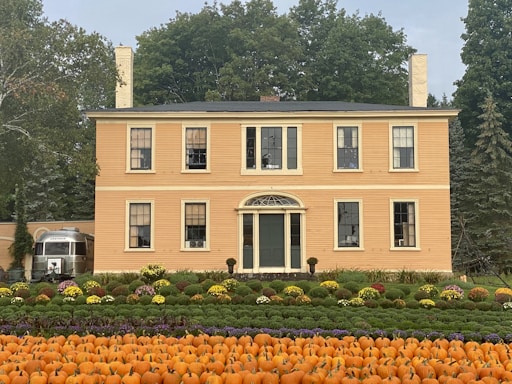 A large, classic yellow house with chimneys and multiple windows stands amidst lush greenery. In front of the house, a garden arranged with rows of orange pumpkins and colorful flowers creates an autumnal scene. On the left side, a silver vintage trailer is partially visible. Tall trees with dense foliage form the backdrop, suggesting a peaceful countryside setting.