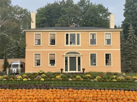 A large, classic yellow house with chimneys and multiple windows stands amidst lush greenery. In front of the house, a garden arranged with rows of orange pumpkins and colorful flowers creates an autumnal scene. On the left side, a silver vintage trailer is partially visible. Tall trees with dense foliage form the backdrop, suggesting a peaceful countryside setting.