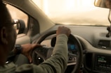 A person with short hair is sitting in the driver's seat of a car, holding the steering wheel with both hands. They are wearing glasses and a watch, and a part of their face is visible. The car's interior is light-colored with visible dashboard features, including an FM radio display and a speedometer showing the speed.