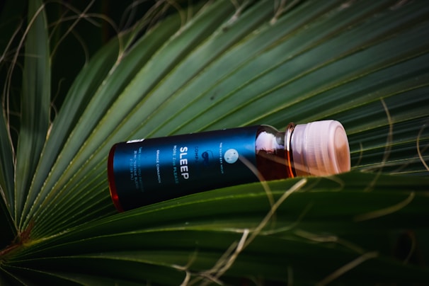 A bottle labeled 'Sleep' is positioned diagonally, placed gently on a large, lush green leaf. The bottle is dark with a blue label and a white cap. The background consists of overlapping palm leaves, creating a natural and tropical setting.