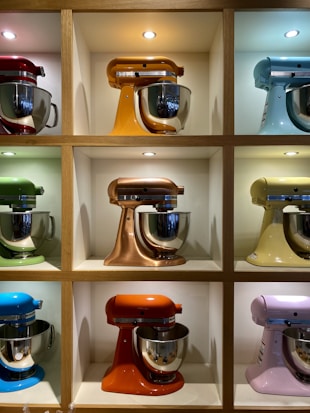 A display of nine colorful stand mixers, each positioned within its own wooden cubby. The mixers come in various colors including red, orange, blue, green, yellow, copper, and pink. Each is fitted with a stainless steel mixing bowl that reflects light from overhead spotlights within each cubby.