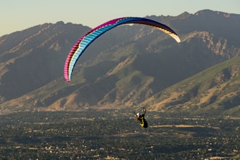 A paraglider is soaring in the air with a vibrant, multicolored canopy. The backdrop features expansive, rugged mountains with green and brown tones. Below, a dense, green forest and rocky terrain stretch out, illuminated by warm sunlight.