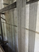 Several vertical steel cables are aligned closely together against a lightly textured concrete wall. The cables have a reflective quality, suggesting they are made of metal. The background includes structural metal beams, indicating an industrial or construction setting.