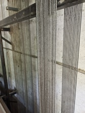 Several vertical steel cables are aligned closely together against a lightly textured concrete wall. The cables have a reflective quality, suggesting they are made of metal. The background includes structural metal beams, indicating an industrial or construction setting.
