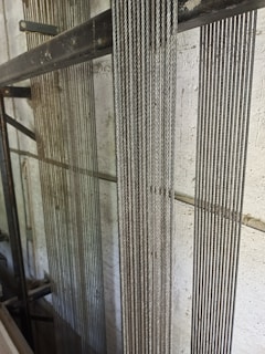 Several vertical steel cables are aligned closely together against a lightly textured concrete wall. The cables have a reflective quality, suggesting they are made of metal. The background includes structural metal beams, indicating an industrial or construction setting.