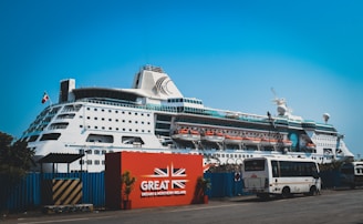 A large white cruise ship docked at a port with multiple decks and lifeboats visible. The ship is positioned behind a red sign with the text 'Great Britain & Northern Ireland' and features a union jack design. A bus and several smaller vehicles are parked near the sign. The sky is clear and blue.
