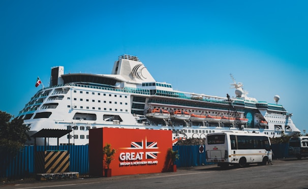 A large white cruise ship docked at a port with multiple decks and lifeboats visible. The ship is positioned behind a red sign with the text 'Great Britain & Northern Ireland' and features a union jack design. A bus and several smaller vehicles are parked near the sign. The sky is clear and blue.