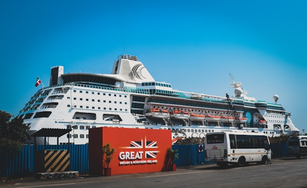 A large white cruise ship docked at a port with multiple decks and lifeboats visible. The ship is positioned behind a red sign with the text 'Great Britain & Northern Ireland' and features a union jack design. A bus and several smaller vehicles are parked near the sign. The sky is clear and blue.