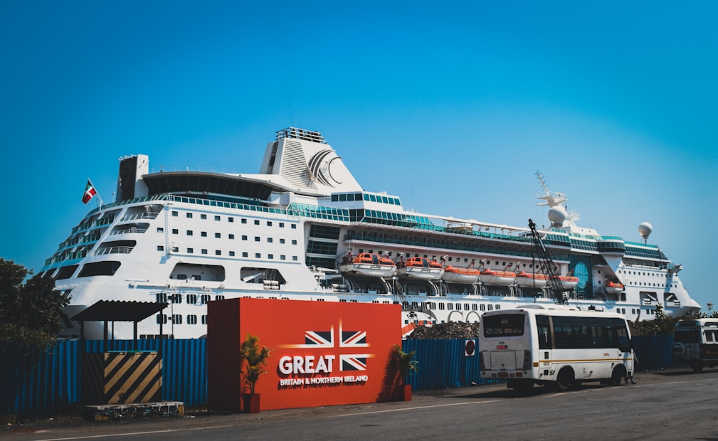 A large white cruise ship docked at a port with multiple decks and lifeboats visible. The ship is positioned behind a red sign with the text 'Great Britain & Northern Ireland' and features a union jack design. A bus and several smaller vehicles are parked near the sign. The sky is clear and blue.