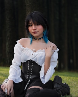 A person with short black hair and blue tips is sitting outdoors against a backdrop of trees. They are wearing a white off-shoulder top with a black corset, a black choker, and black boots. The outfit has a gothic or alternative style.