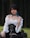A person with short black hair and blue tips is sitting outdoors against a backdrop of trees. They are wearing a white off-shoulder top with a black corset, a black choker, and black boots. The outfit has a gothic or alternative style.