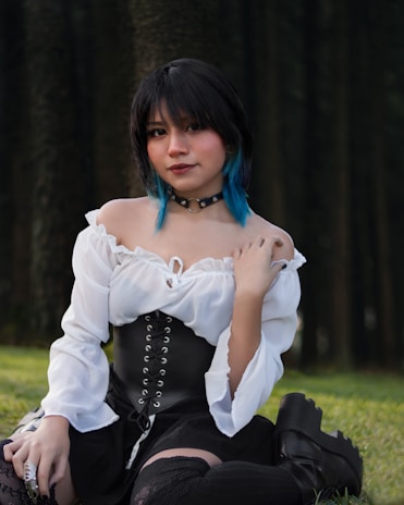 A person with short black hair and blue tips is sitting outdoors against a backdrop of trees. They are wearing a white off-shoulder top with a black corset, a black choker, and black boots. The outfit has a gothic or alternative style.