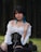 A person with short black hair and blue tips is sitting outdoors against a backdrop of trees. They are wearing a white off-shoulder top with a black corset, a black choker, and black boots. The outfit has a gothic or alternative style.