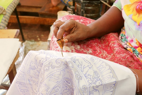 A person is using a small tool to apply a design onto fabric. The fabric has intricate blue patterns, and the person is holding a brass tool in one hand. The setting appears to be a workspace or a craft area, with wooden surfaces and textiles visible.