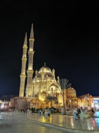 A grand mosque with two tall minarets and multiple domes, illuminated in warm golden lights against a dark night sky. The architecture is intricate, featuring arches and ornate patterns. People stroll along the paved area, some stopping to admire the view. The scene has a lively atmosphere with palm trees and surrounding buildings adding to the vibrant setting.