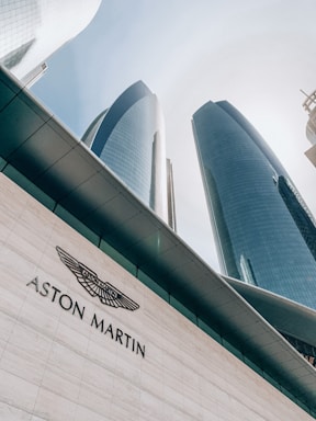 Tall modern skyscrapers with reflective glass facades rise against a clear sky. The foreground features a building with the Aston Martin logo prominently displayed on a white tiled surface.