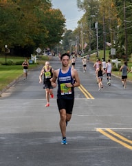 A group of runners participating in a road race on a tree-lined street during the day. The road is flanked by green lawns and utility poles. Some runners wear bib numbers, and one in front is wearing a blue sleeveless shirt.