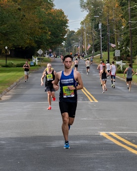 A group of runners participating in a road race on a tree-lined street during the day. The road is flanked by green lawns and utility poles. Some runners wear bib numbers, and one in front is wearing a blue sleeveless shirt.