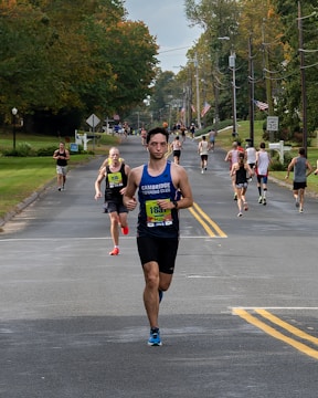 A group of runners participating in a road race on a tree-lined street during the day. The road is flanked by green lawns and utility poles. Some runners wear bib numbers, and one in front is wearing a blue sleeveless shirt.