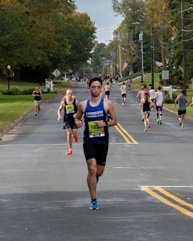 A group of runners participating in a road race on a tree-lined street during the day. The road is flanked by green lawns and utility poles. Some runners wear bib numbers, and one in front is wearing a blue sleeveless shirt.