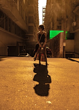 A person with dreadlocks stands in an urban alleyway, casting a long shadow on the pavement. The scene is lit with a warm, golden hue from the sunlight filtering between tall buildings. A hint of greenery appears on a wall in the background, contrasting against the predominantly brown and yellow tones.
