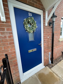 A vibrant blue door with the number 10 displayed at the top. A lush green wreath is hung on the door, adding a touch of nature. The door features a mail slot and a handle with a lock. Surrounding the door is a red brick wall, and to the right, a black lantern is mounted. Light stone tiles form the pathway leading to the entrance.
