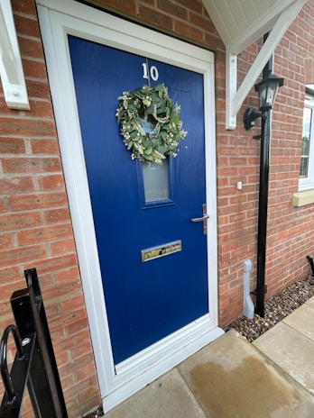 A vibrant blue door with the number 10 displayed at the top. A lush green wreath is hung on the door, adding a touch of nature. The door features a mail slot and a handle with a lock. Surrounding the door is a red brick wall, and to the right, a black lantern is mounted. Light stone tiles form the pathway leading to the entrance.