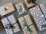 A collection of gift-wrapped boxes with brown paper and various decorations such as ribbons, pinecones, and green leaves. Some of the wrapping paper is printed with white tree patterns. Tags are attached to several packages, and the arrangement is on a textured surface.