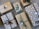 A collection of gift-wrapped boxes with brown paper and various decorations such as ribbons, pinecones, and green leaves. Some of the wrapping paper is printed with white tree patterns. Tags are attached to several packages, and the arrangement is on a textured surface.