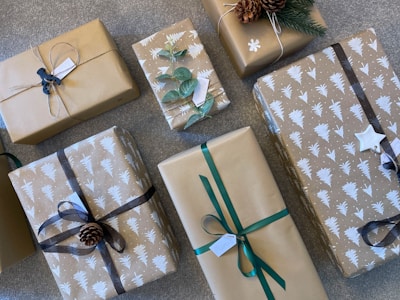 A collection of gift-wrapped boxes with brown paper and various decorations such as ribbons, pinecones, and green leaves. Some of the wrapping paper is printed with white tree patterns. Tags are attached to several packages, and the arrangement is on a textured surface.