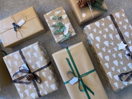 A collection of gift-wrapped boxes with brown paper and various decorations such as ribbons, pinecones, and green leaves. Some of the wrapping paper is printed with white tree patterns. Tags are attached to several packages, and the arrangement is on a textured surface.