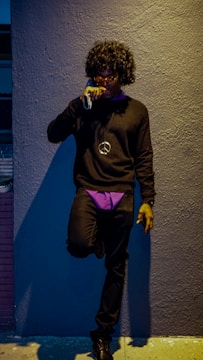 A person wearing a curly wig, sunglasses, and clothing in shades of black and purple stands against a textured wall. They hold a cigarette in their hand and have a peace sign necklace.