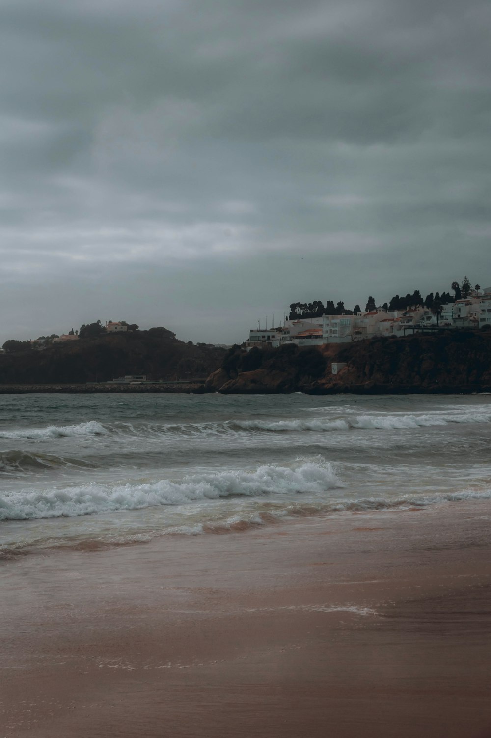 a beach with waves coming in to shore and houses on a hill in the distance