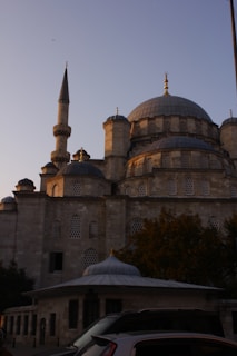 A historic mosque with a prominent dome and tall minaret constructed from light-colored stone. The structure features intricate lattice windows and decorative golden finials. The building is partially surrounded by trees, and there are vehicles parked nearby. The sky is clear with a hint of warm sunlight illuminating the facade.