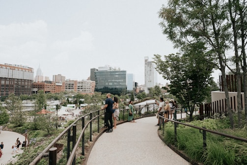 A landscaped urban park pathway lined with trees and greenery, bustling with people walking and socializing. Modern buildings form the backdrop with a mix of glass and brick facades. The scene captures a mix of nature and city elements, with groups of people enjoying the outdoors.
