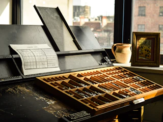A vintage typesetting case filled with metal type pieces sits on a table. Next to it is a slanted wooden desk with a typographic chart pinned on it. On the windowsill, there is a ceramic pitcher and a framed artwork, with sunlight streaming through the window casting warm shadows.